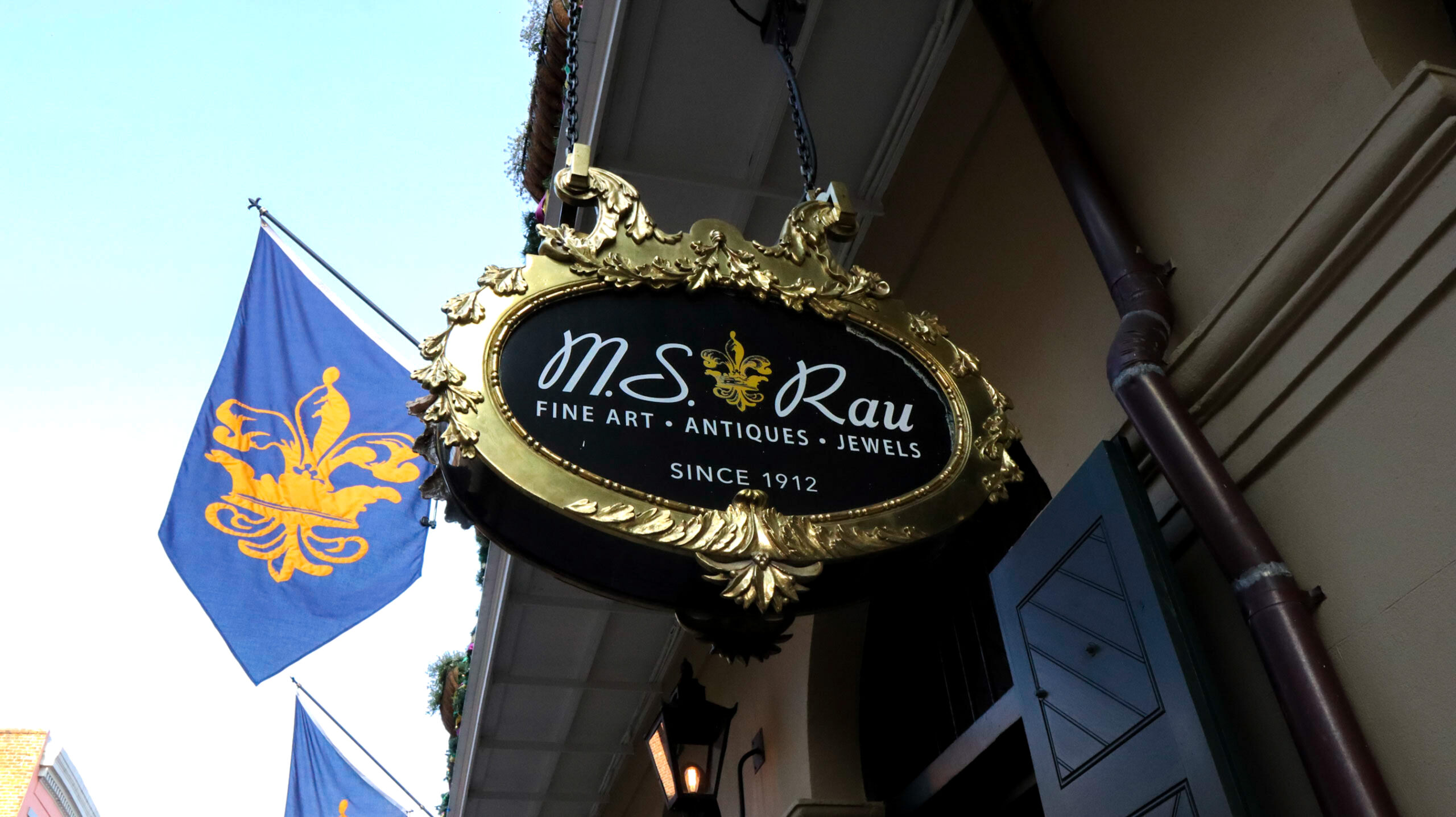 Exterior sign of MS Rau Gallery in New Orleans with decorative gold frame and fleur-de-lis flags.
